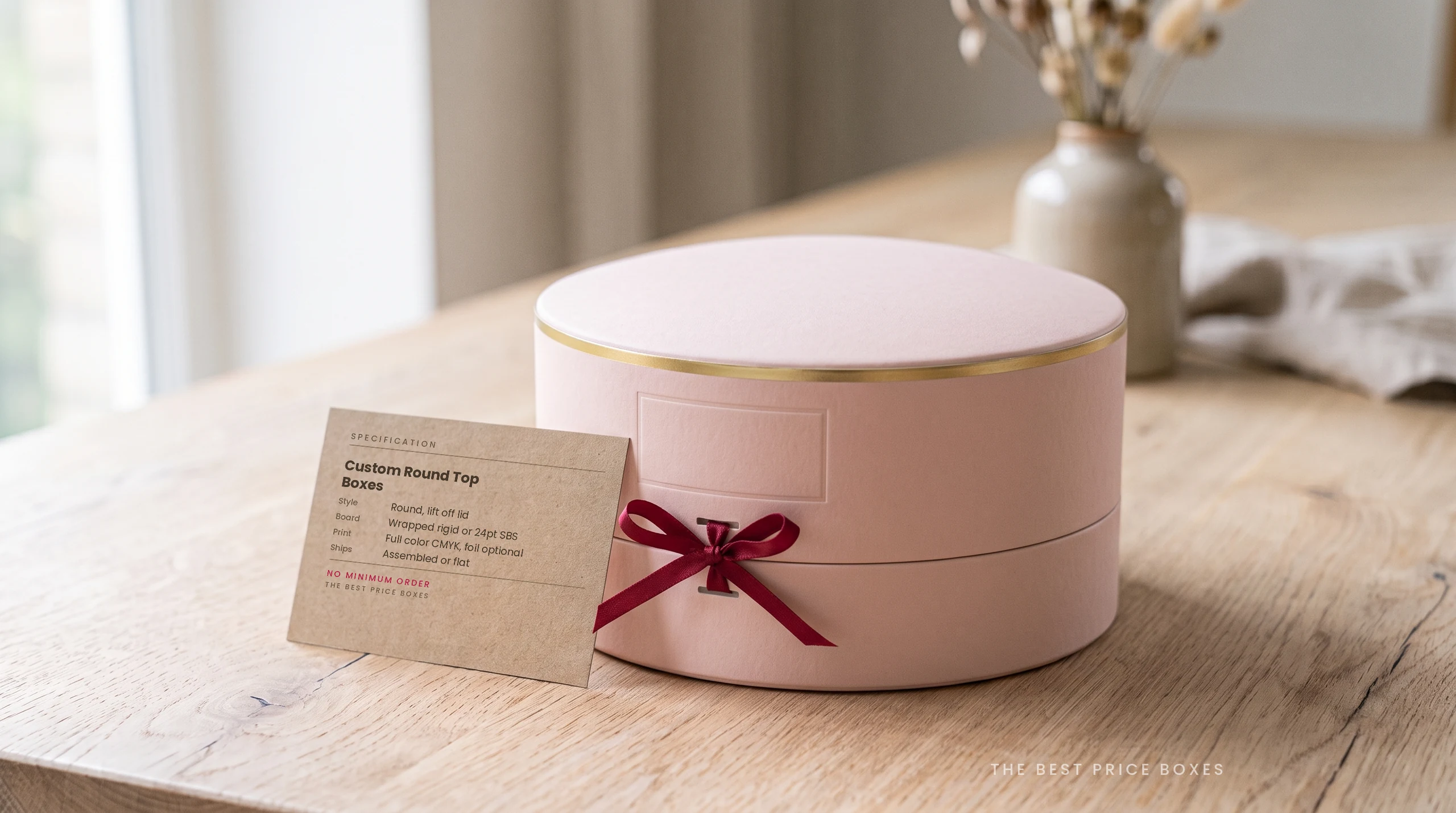 Custom round top box in blush pink with a gold band and lift off domed lid, next to a kraft spec card listing board, print and shipping options.
